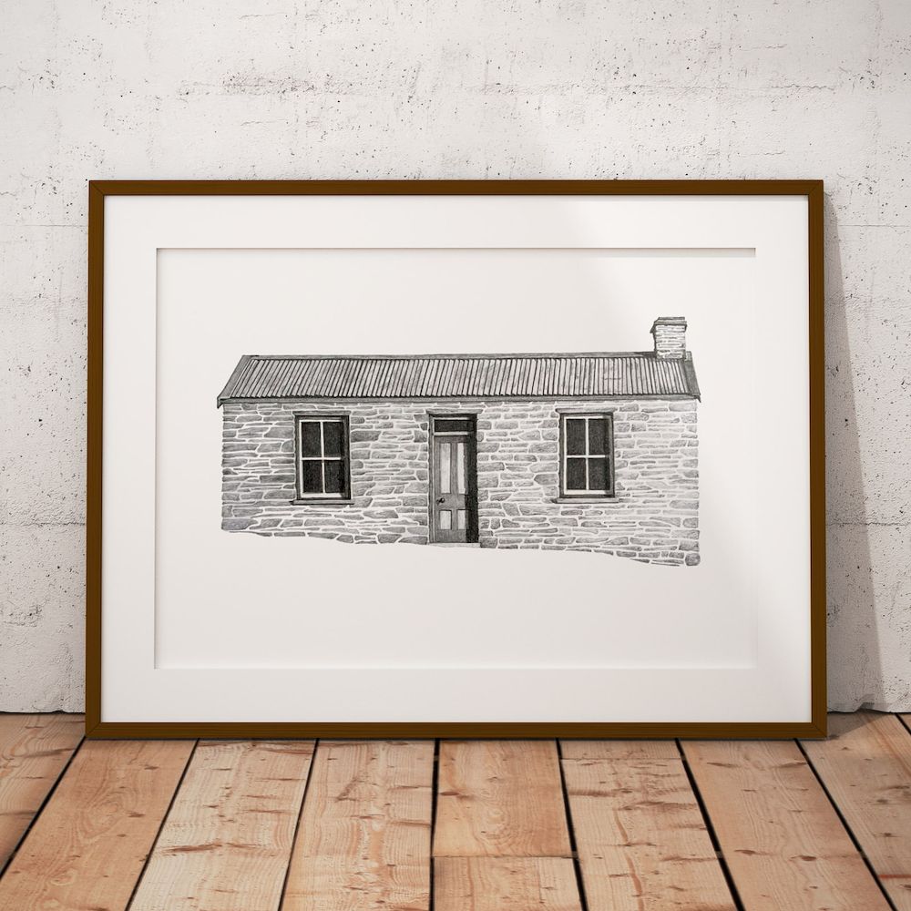 The Mitchells cottage print in a slim wooden frame with a white mount, leaning against a concrete wall